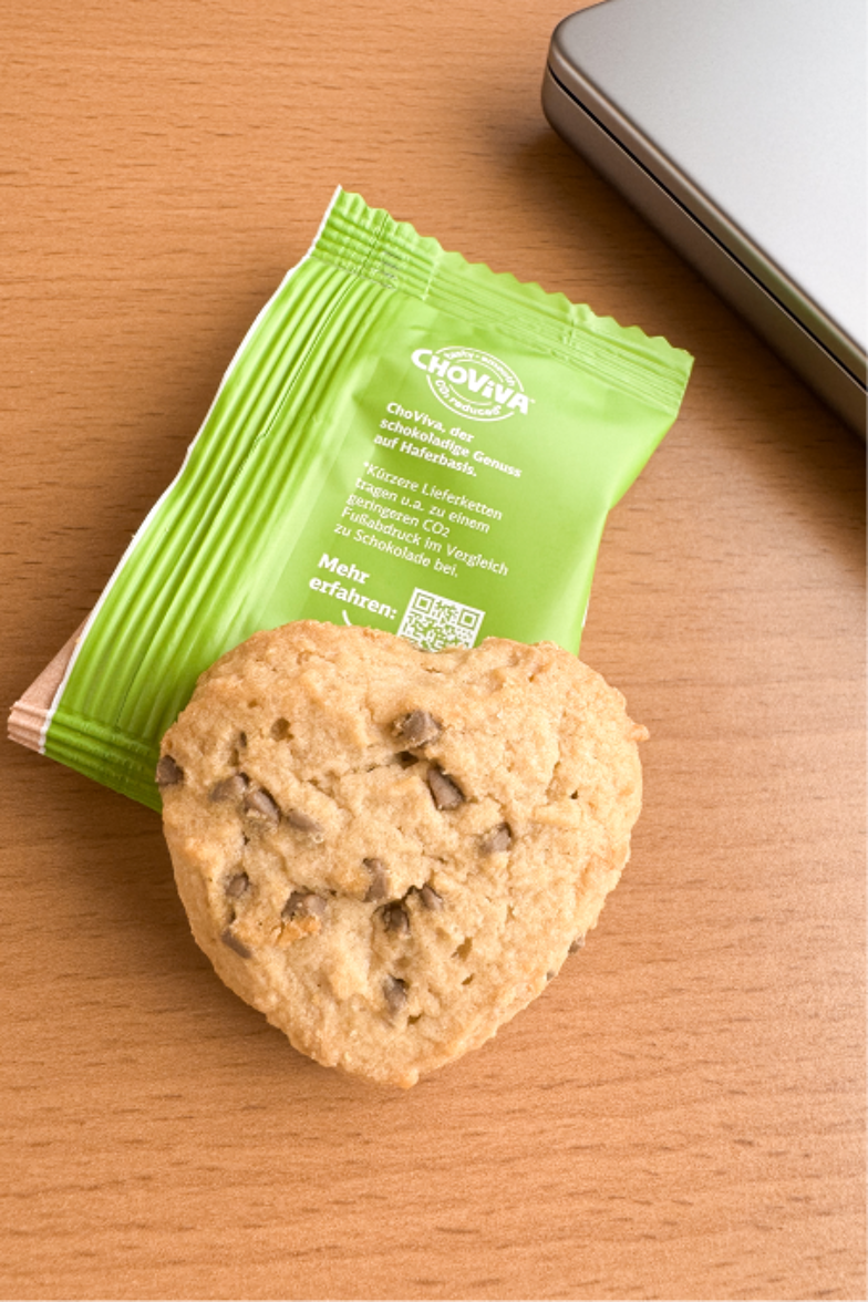 A heart-shaped cookie lies on a green package with the inscription 'Choviva' on a wooden table next to a laptop.